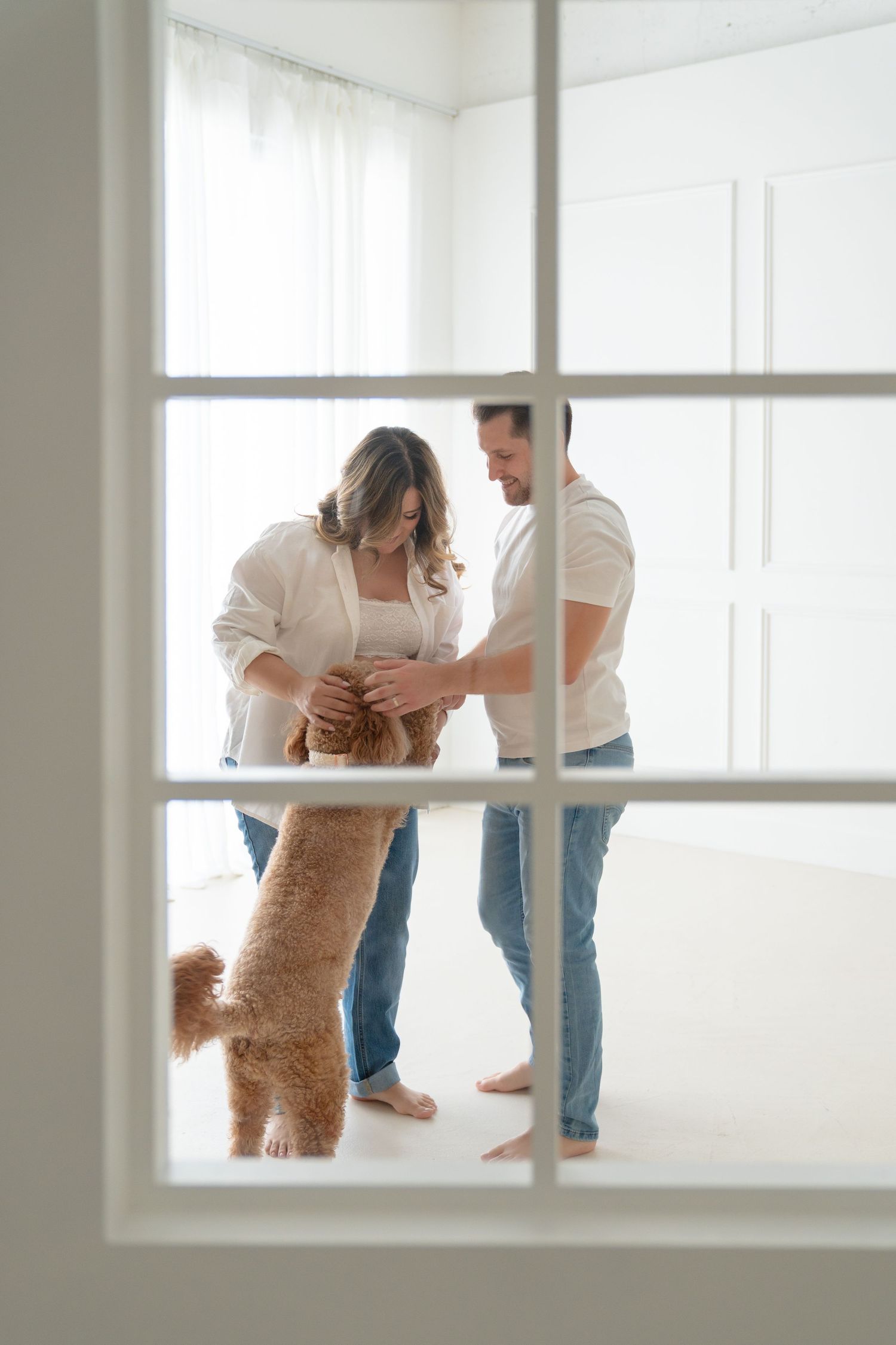 Couple in white room playing with golden doodle dog, viewed through window panes.