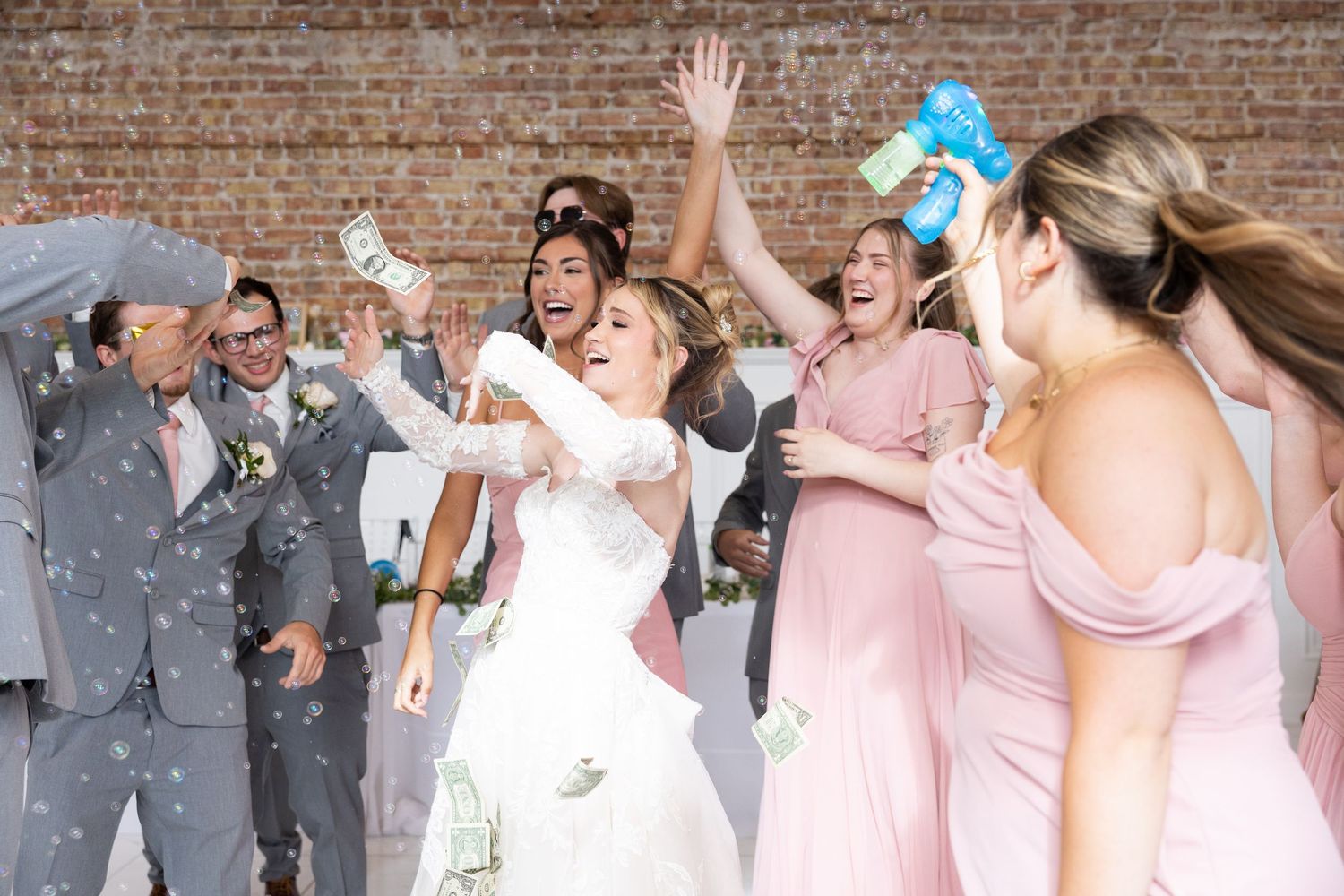 Bride and bridesmaids in pink dresses celebrate joyfully with confetti at a wedding reception near a brick wall.