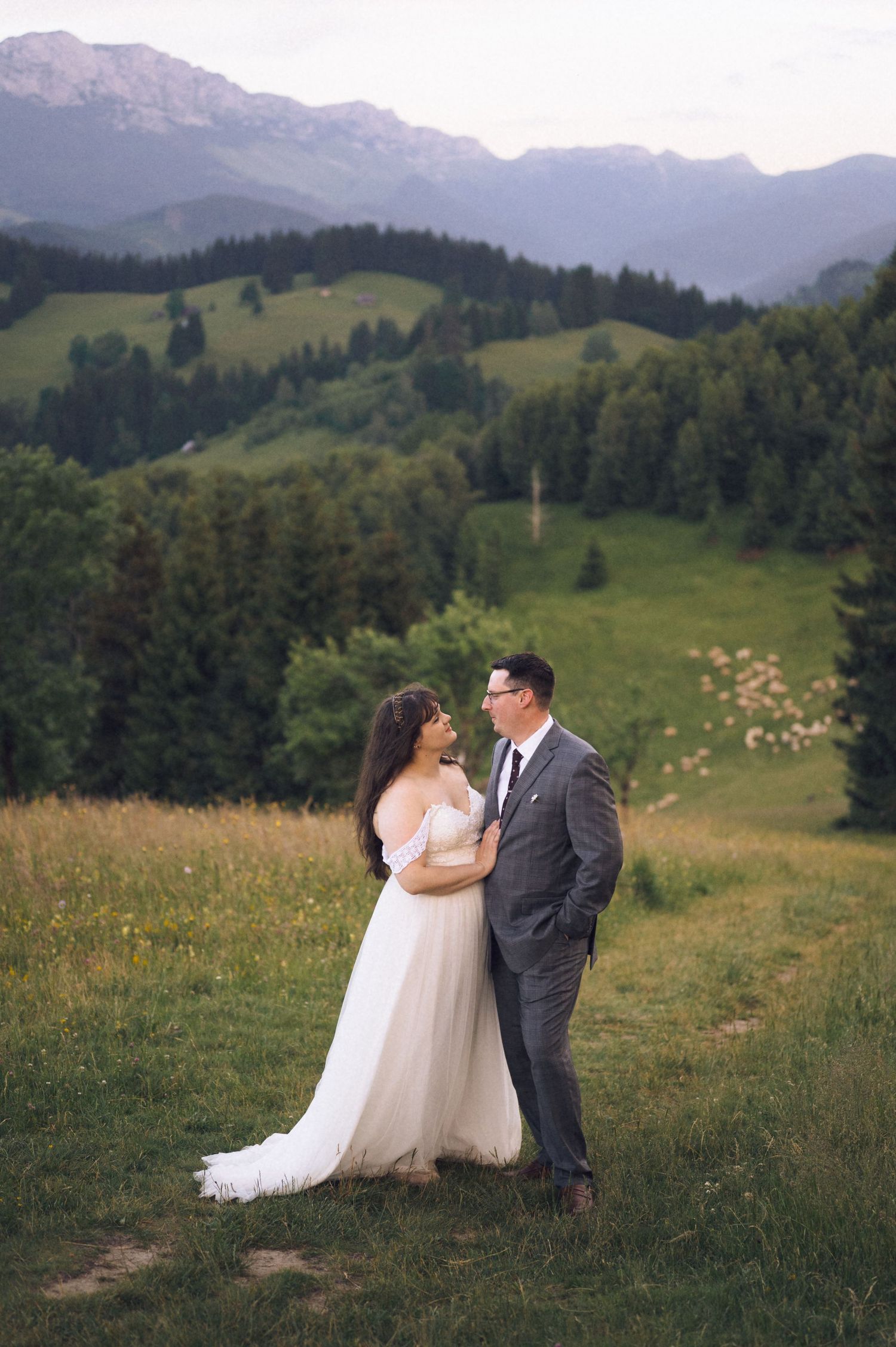 Bride in white gown and groom in gray suit stand in alpine meadow with mountain forest backdrop at dusk.