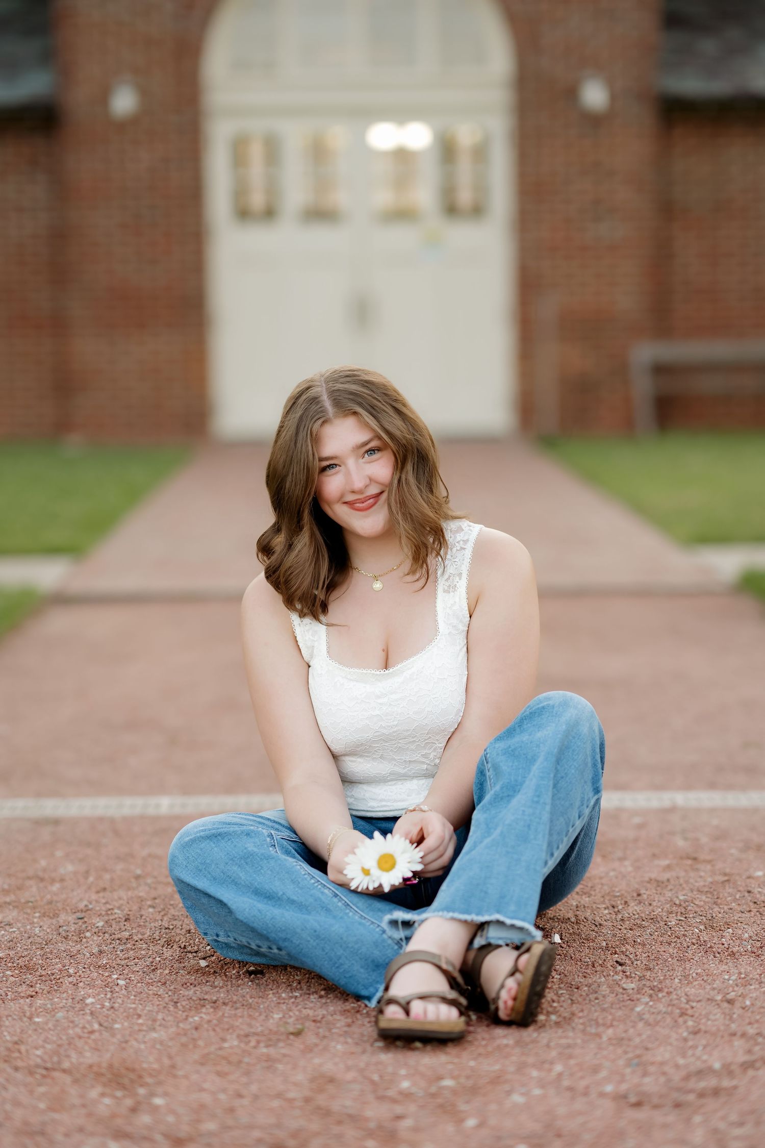 Young woman sitting cross-legged on brick path holding a daisy, smiling in white top and jeans near red brick building.