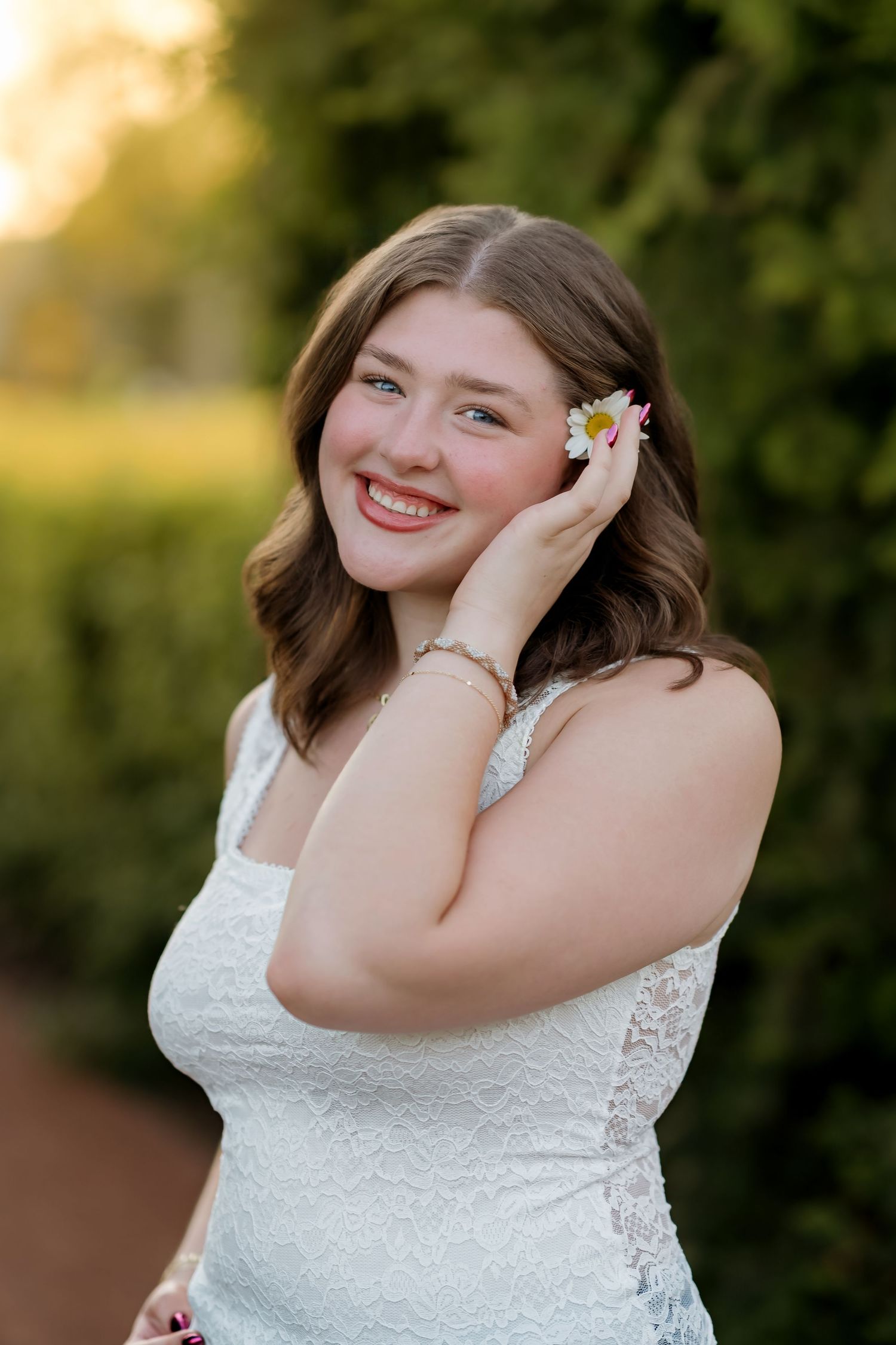 Young woman in white lace dress holding a daisy flower near her face, smiling outdoors in golden hour light.