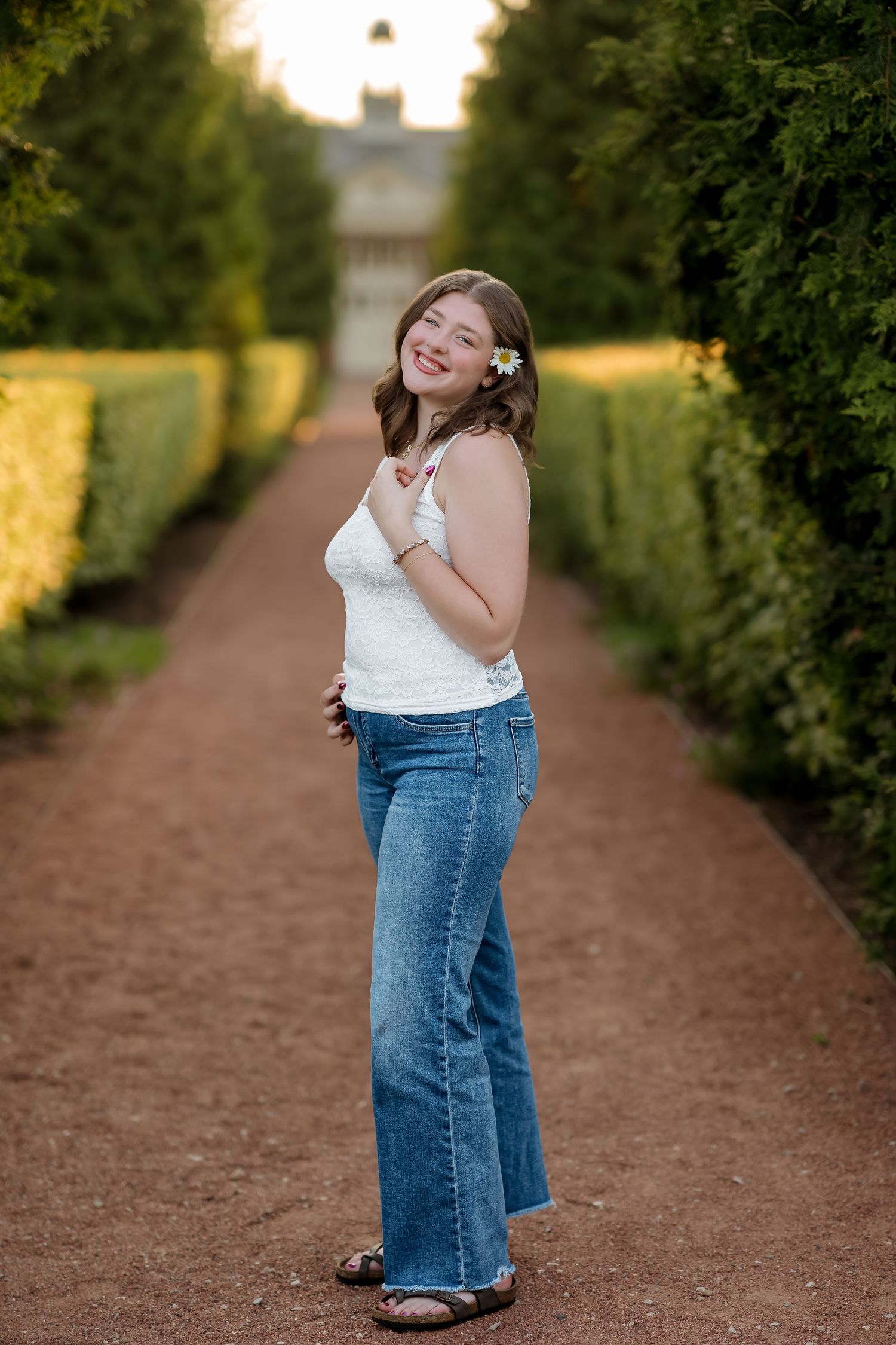 Smiling young woman in white top and jeans poses on a garden path lined with tall green hedges.