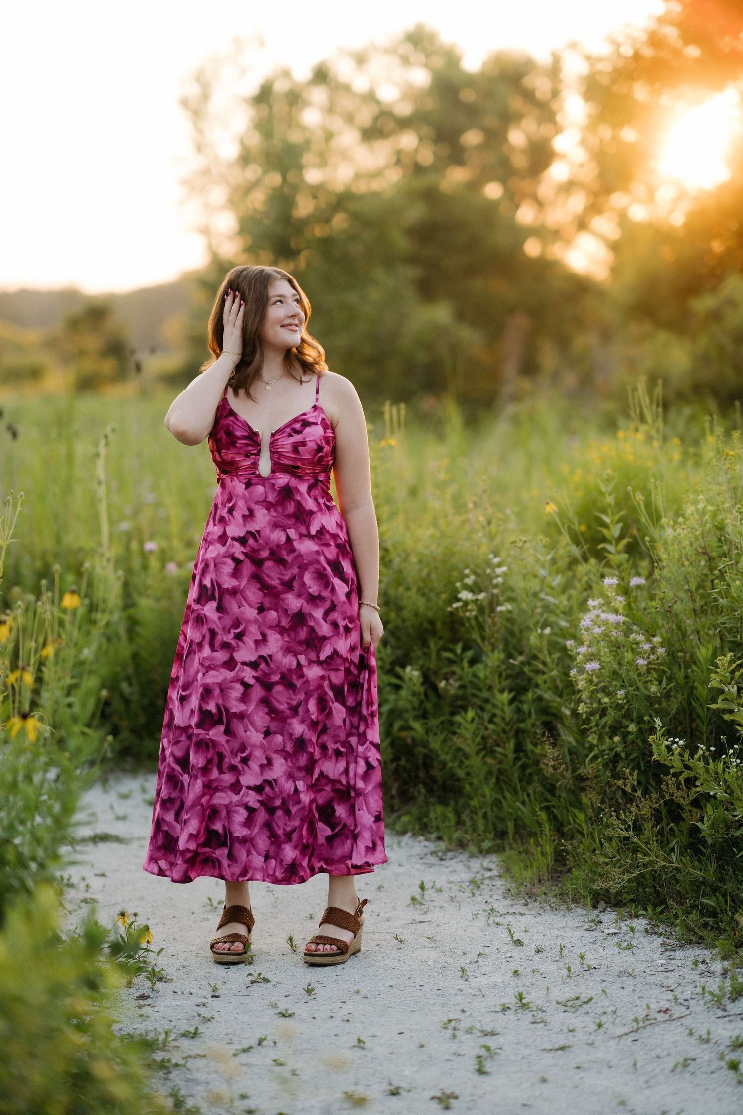 Young woman in pink floral midi dress smiling on a garden path at golden hour sunset.