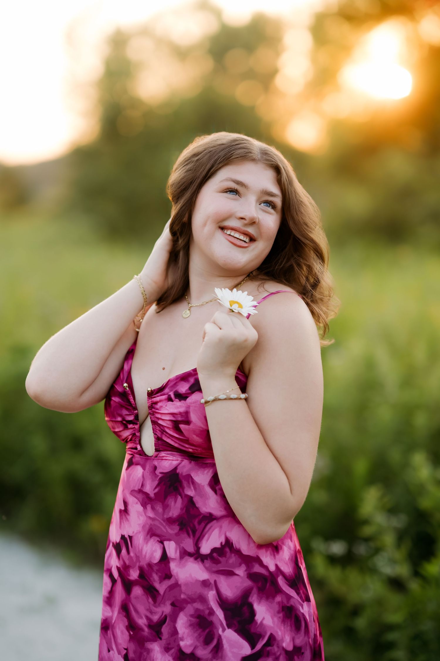 Young woman in pink floral dress holding a daisy, smiling outdoors at golden hour sunset.