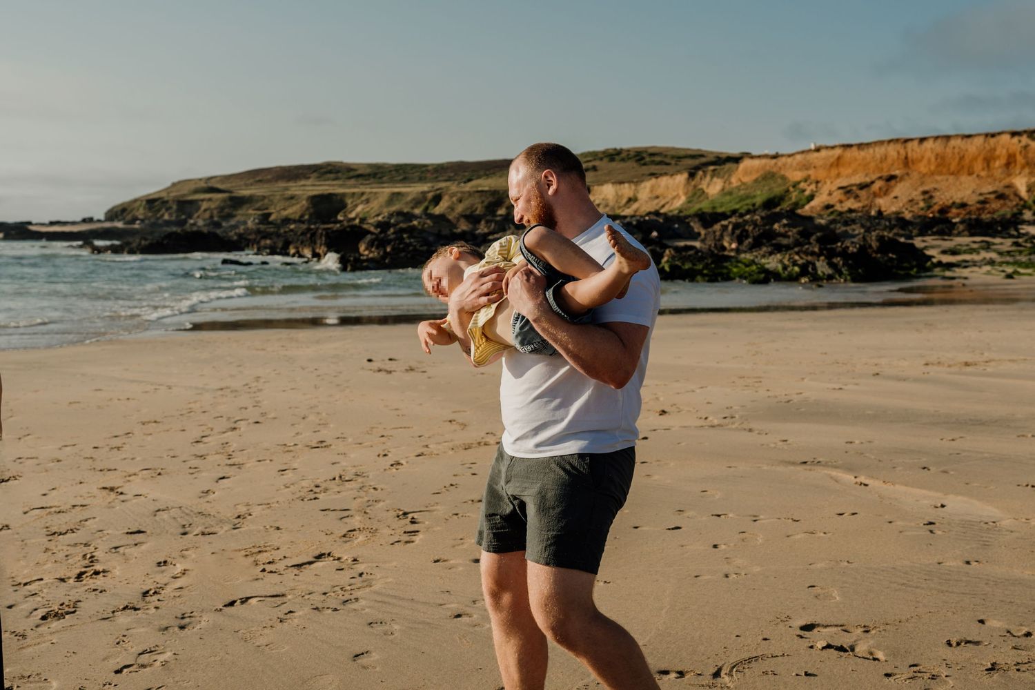 Father holding baby on sandy beach with rocky cliffs and ocean in background on sunny day.