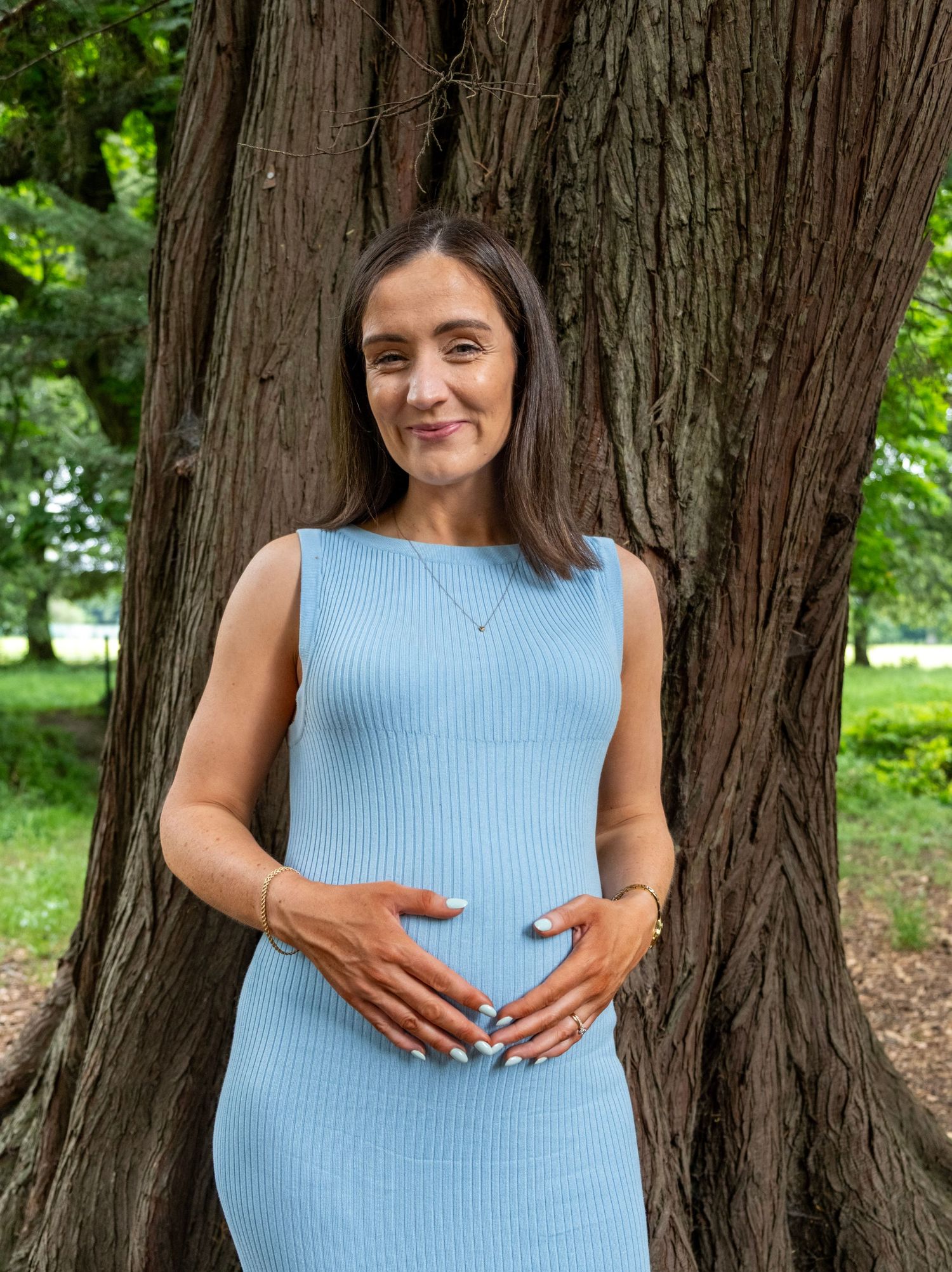 Pregnant woman in light blue ribbed dress posing by large tree trunk in lush green park setting.
