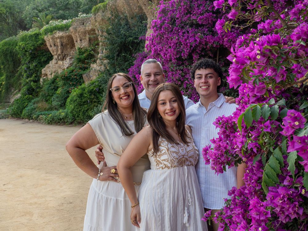 Family of four posing beside vibrant purple bougainvillea flowers outdoors, smiling together in white and light-colored outfits.