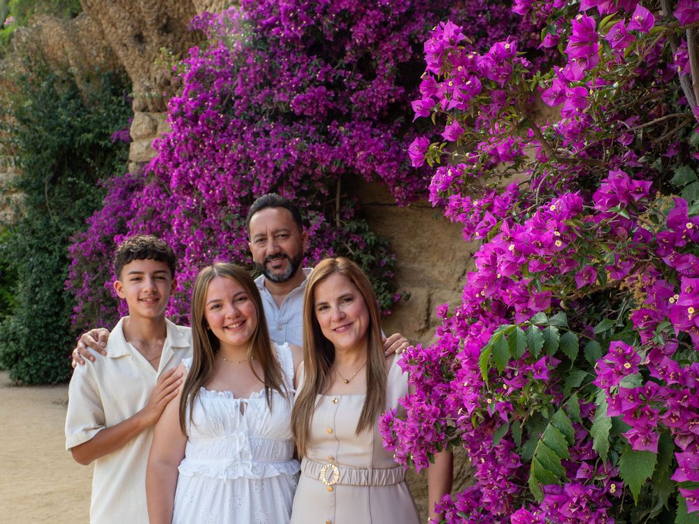 Family of four smiling in front of vibrant purple bougainvillea flowers in a sunny outdoor setting.