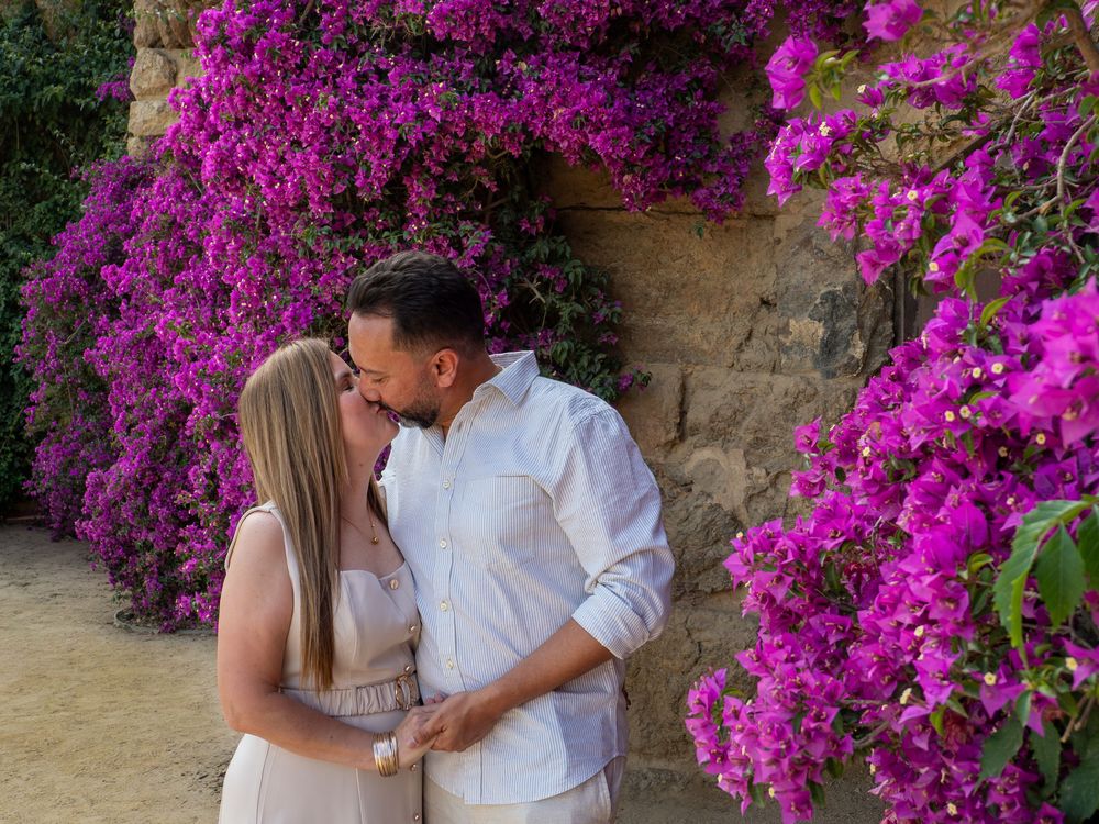 Couple sharing a romantic kiss surrounded by vibrant purple bougainvillea flowers against a stone wall.
