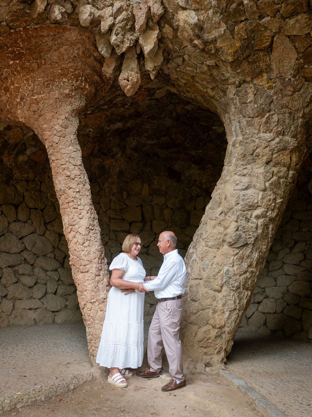 Elderly couple holding hands under stone arches at Park Güell, Barcelona, wearing white and beige outfits.
