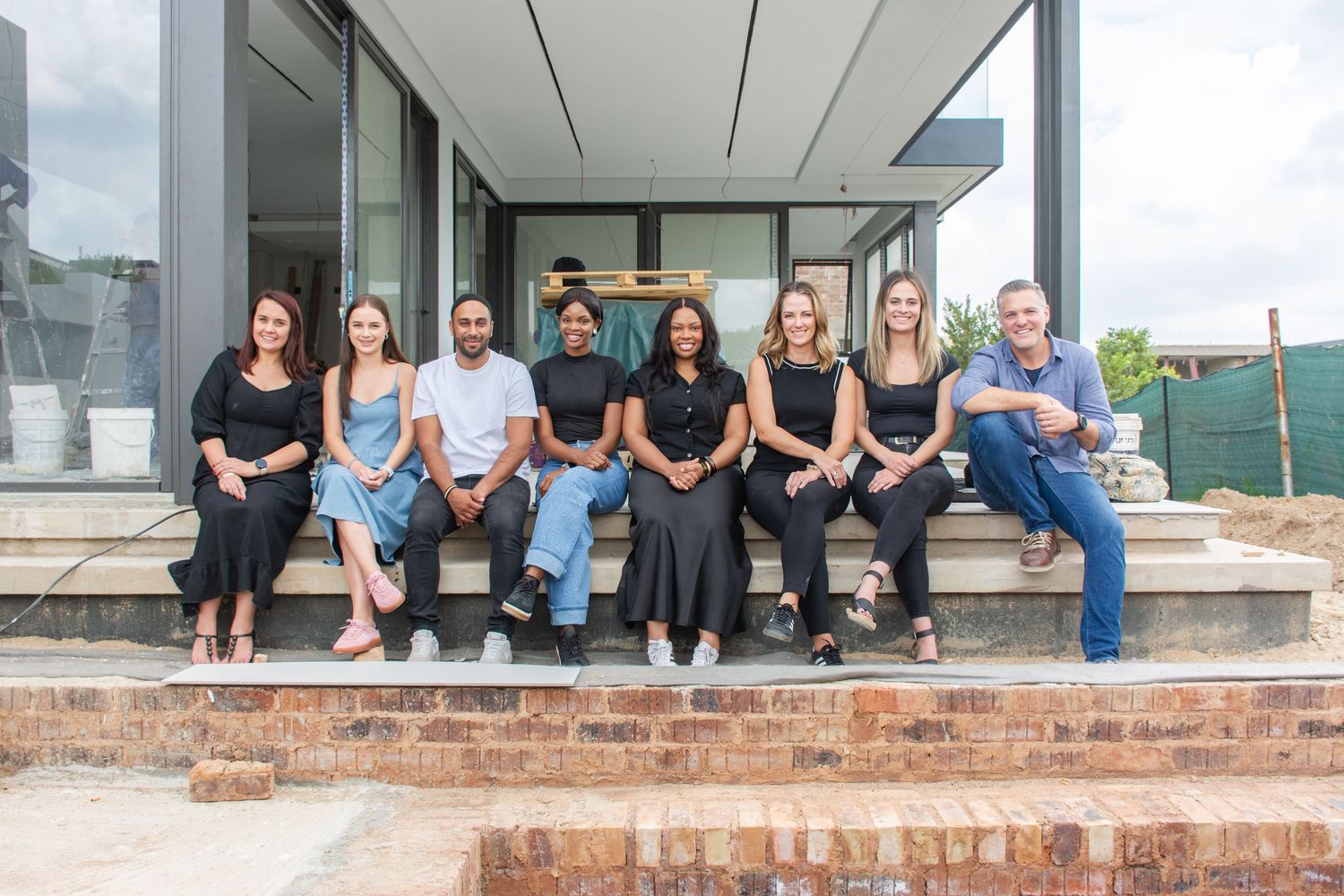 A diverse team of seven interior design professionals smiling together on outdoor steps of a modern building.