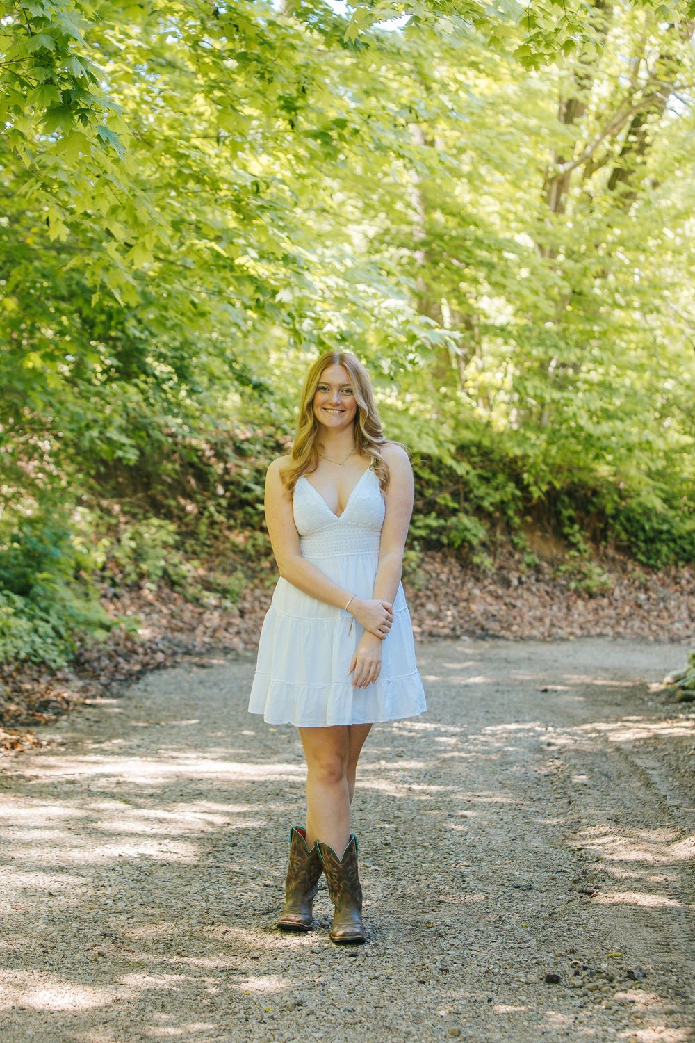 Young woman in white mini dress and cowboy boots smiling on a sunlit gravel path surrounded by lush green trees.