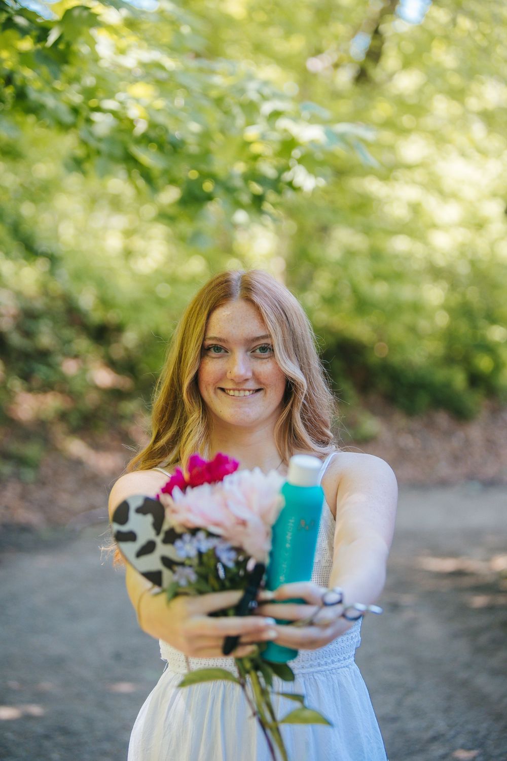 Smiling young woman in white dress holding pink flowers and blue bottle outdoors in sunlit forest path.