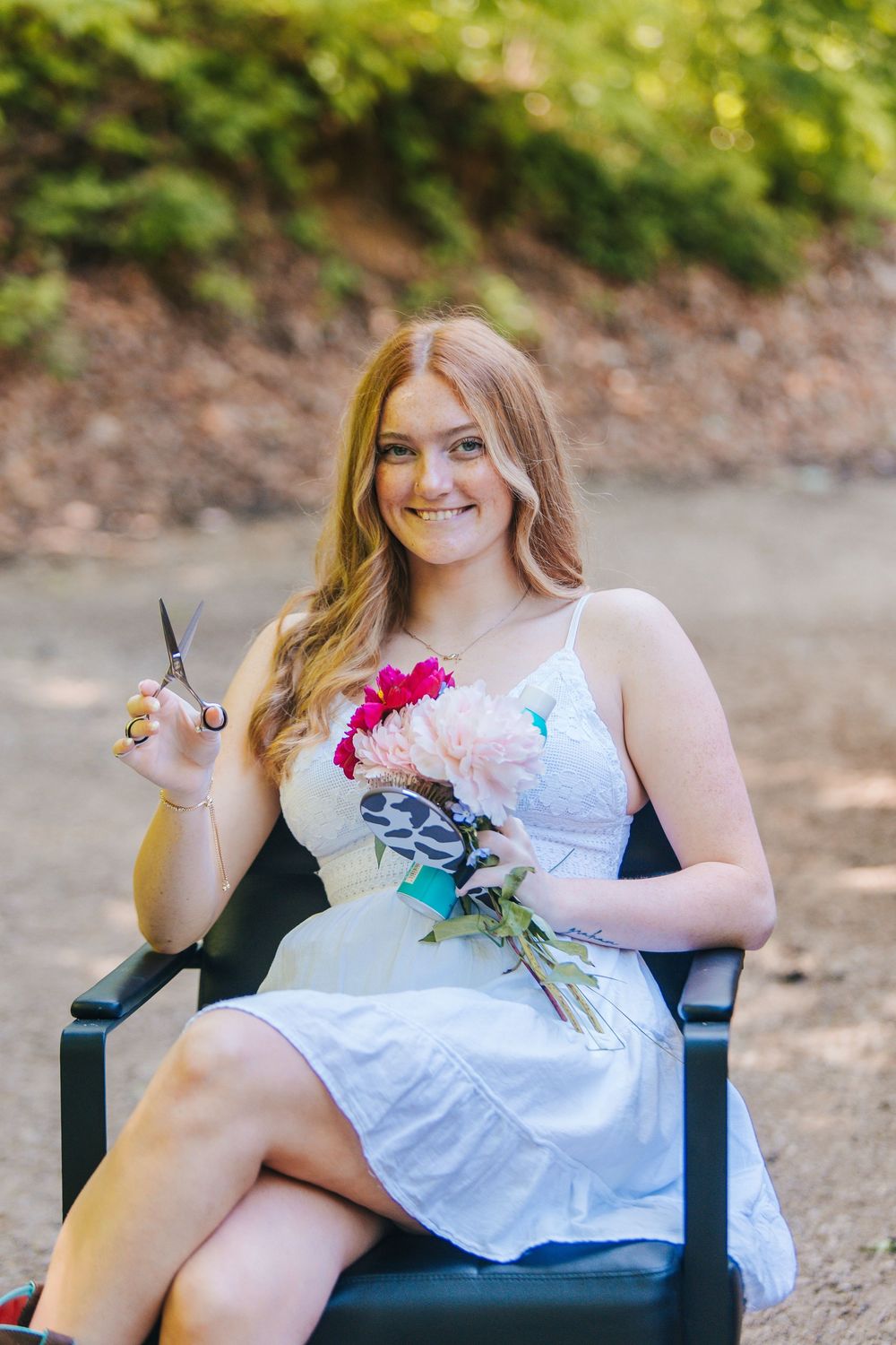 Smiling young woman in white dress holding flowers and scissors, sitting outdoors in a garden setting.