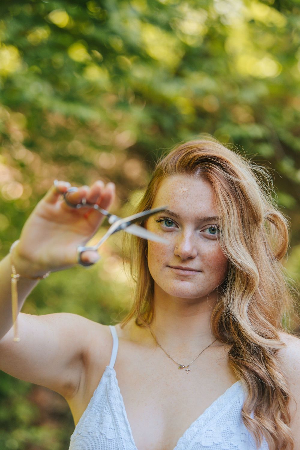 Young woman with wavy red hair holds scissors near her face outdoors, wearing white lace top with glitter makeup.