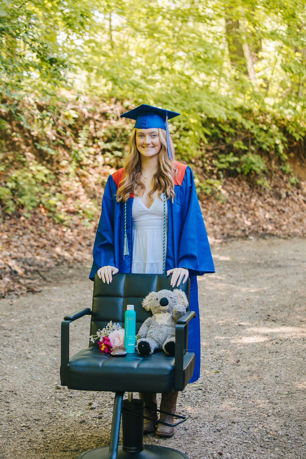 Young woman in blue graduation cap and gown poses outdoors with a chair holding a teddy bear and flowers.