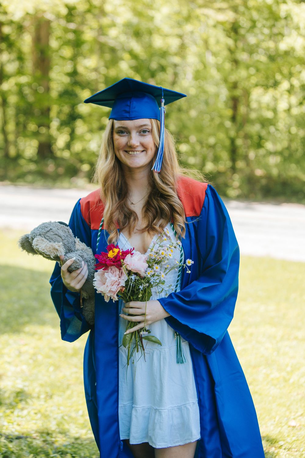 Smiling graduate in blue cap and gown holds flowers and teddy bear outdoors on a sunny day.