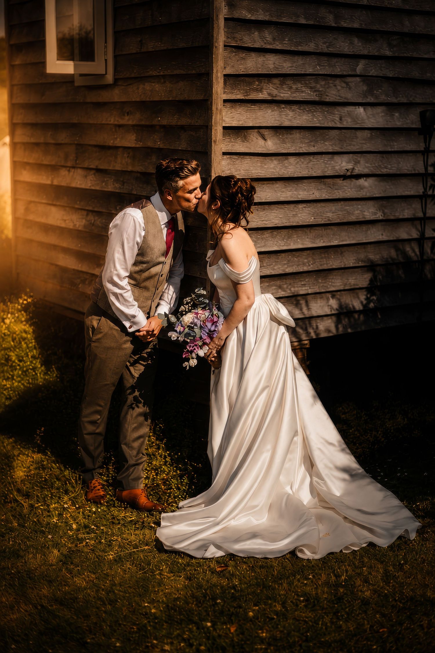 Bride and groom share a golden hour kiss beside a rustic barn, bride holding a wildflower bouquet in a flowing white gown.