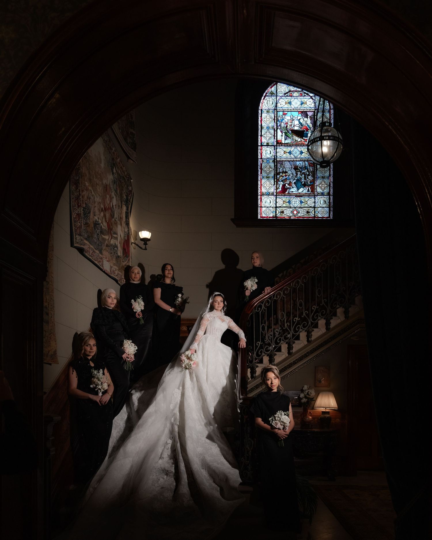 Bride in white gown with bridesmaids in black dresses on dramatic castle staircase.