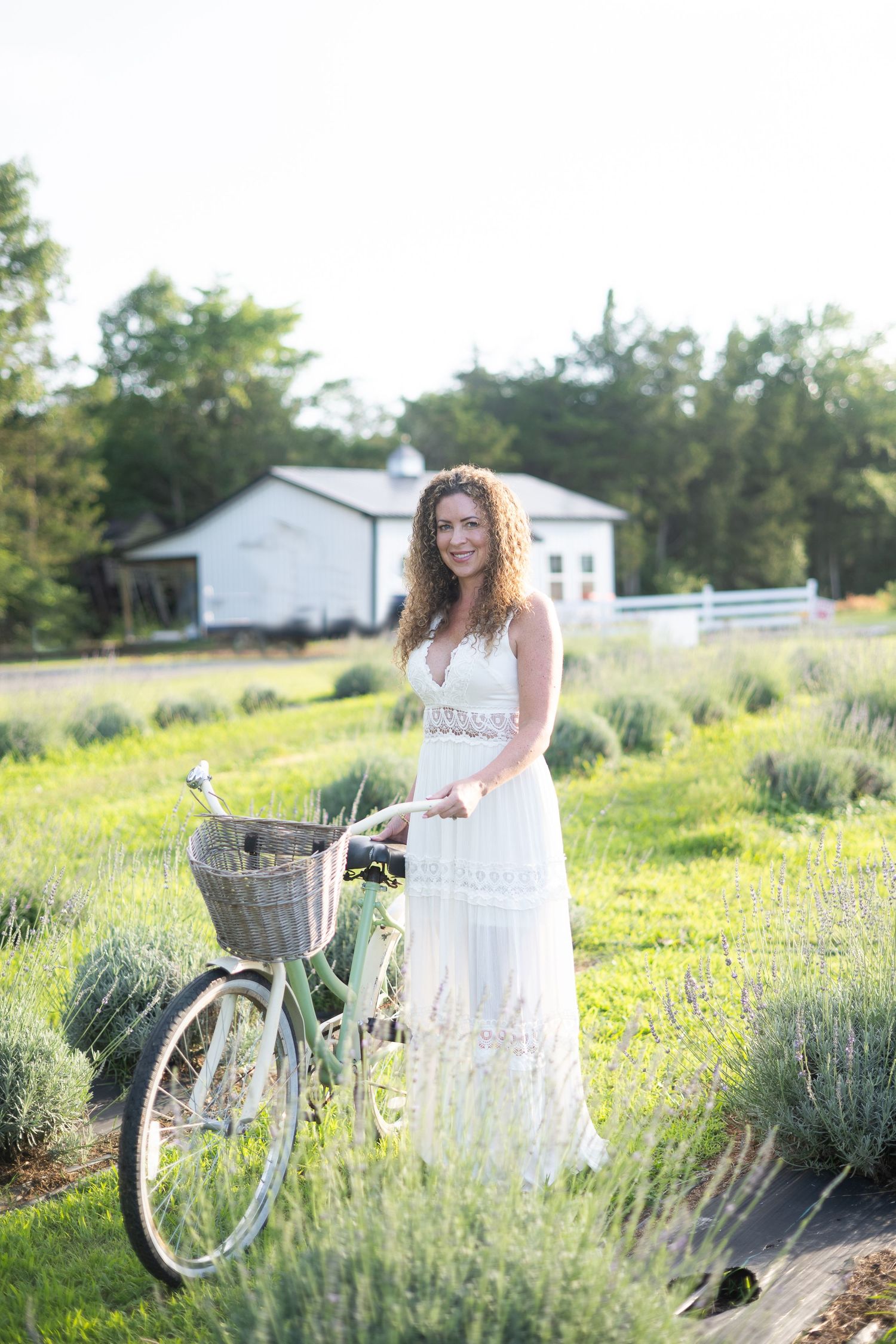 Woman in white dress posing with vintage bicycle in lavender field near white barn on sunny day.