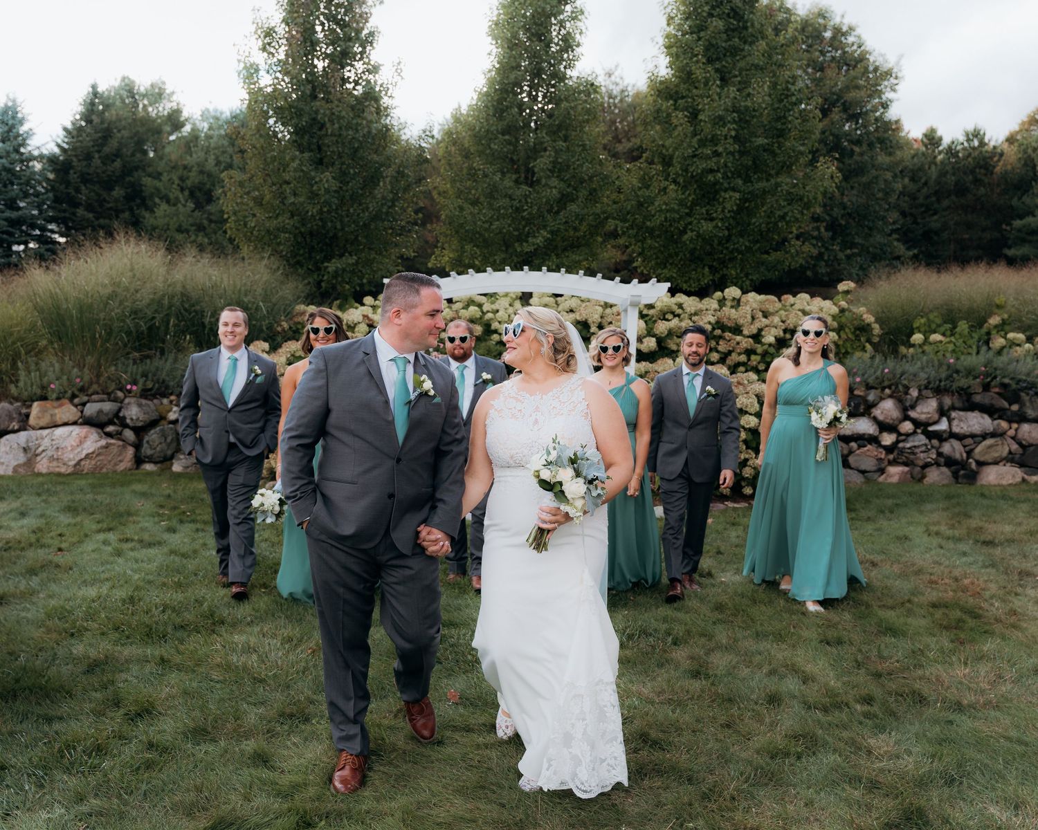 A bride and groom walk hand-in-hand outdoors, with wedding party in teal and gray attire behind them.