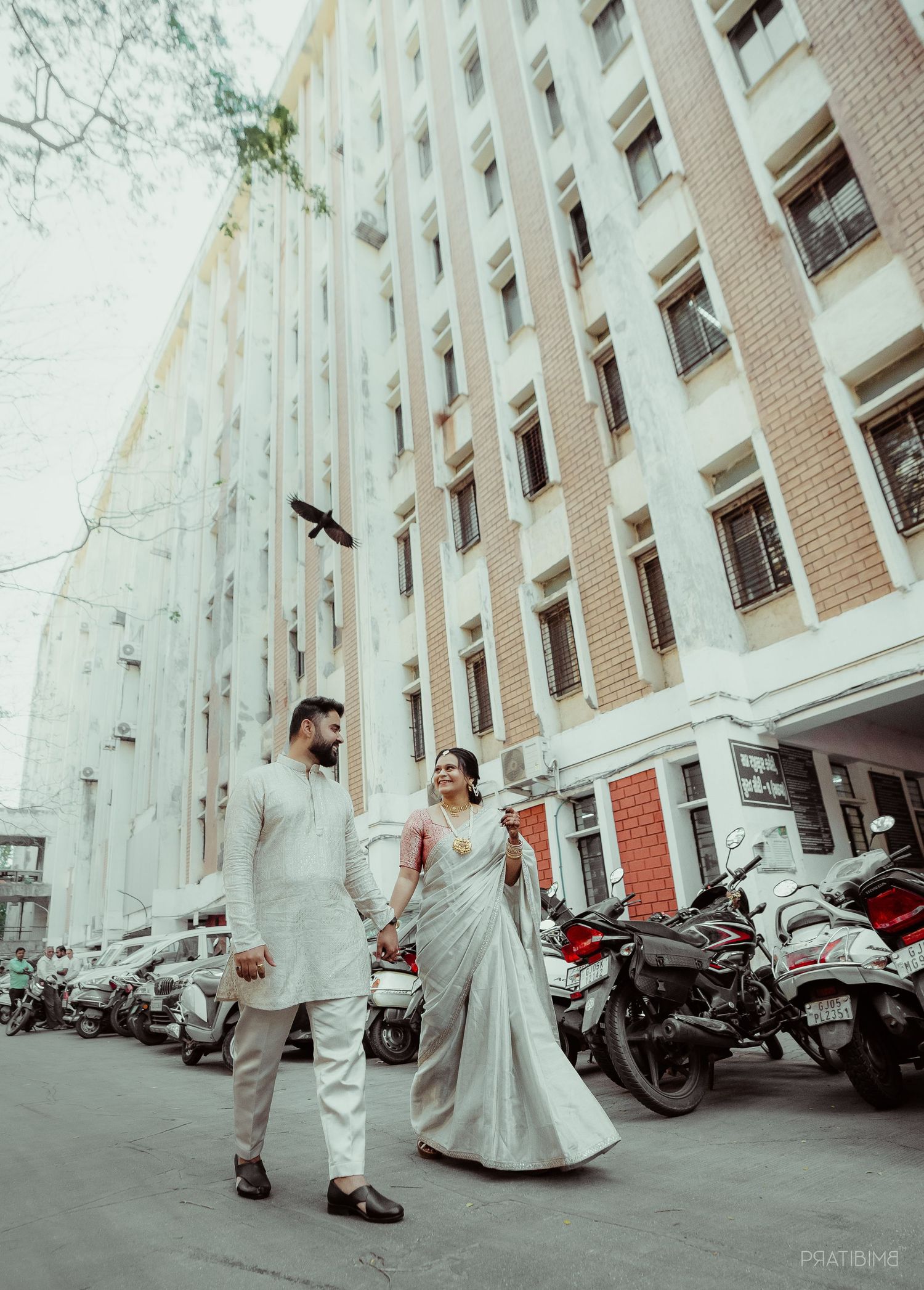 Indian couple in traditional wedding attire walking hand in hand on a city street with motorcycles and a multi-story building.