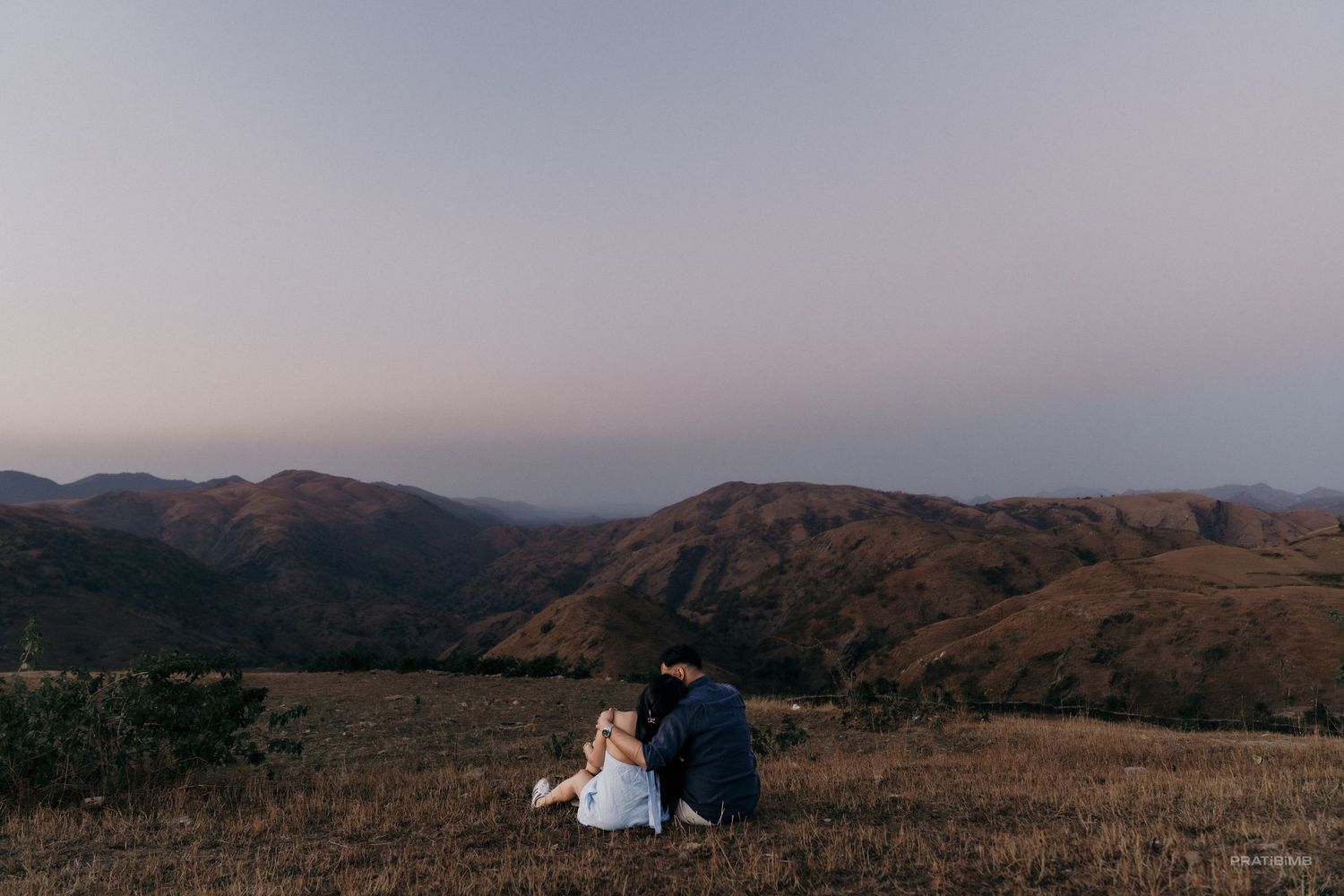 Couple sitting together on a hilltop at dusk overlooking a scenic mountain valley landscape.