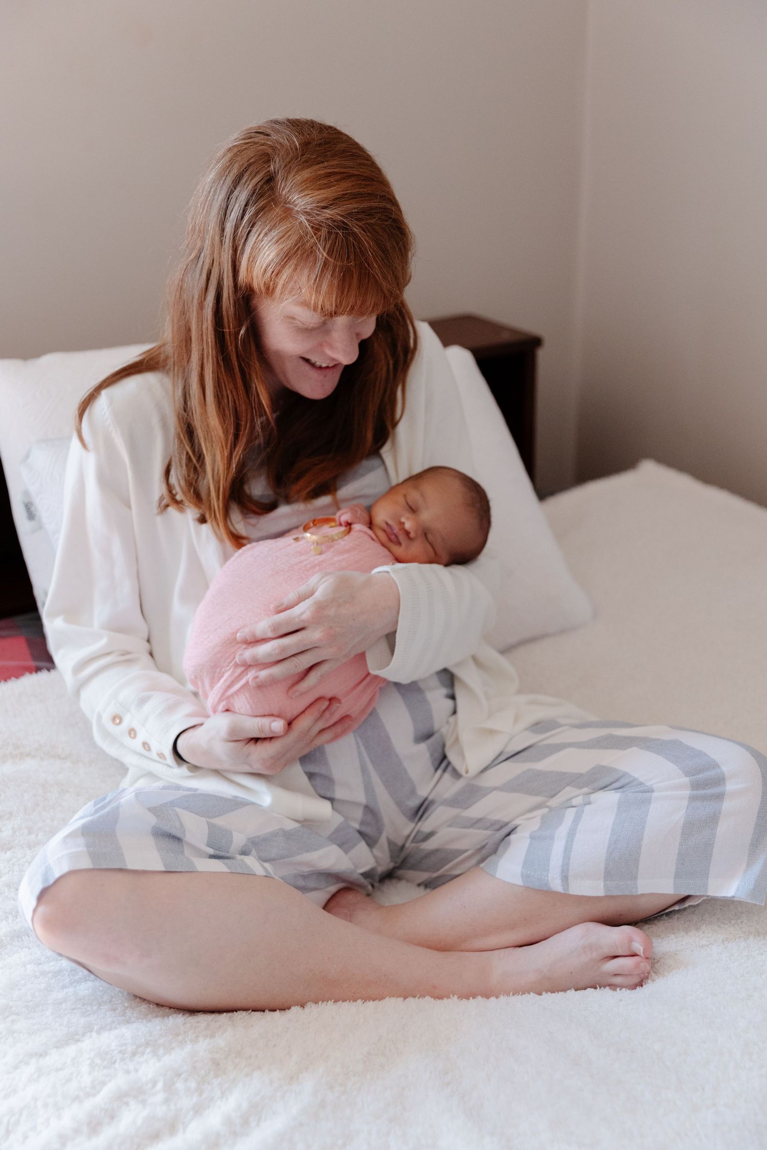 Red-haired mother in striped pajamas sits cross-legged on bed, cradling sleeping newborn baby wrapped in pink blanket.