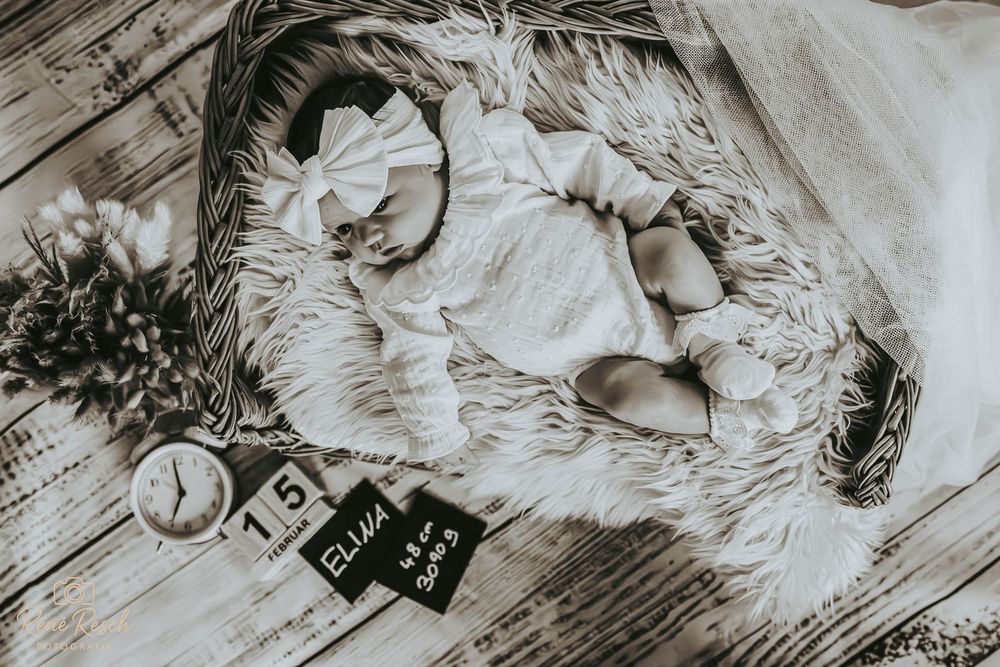 Black and white newborn photo of baby Ellie in a wicker basket with fur, clock, and milestone blocks.