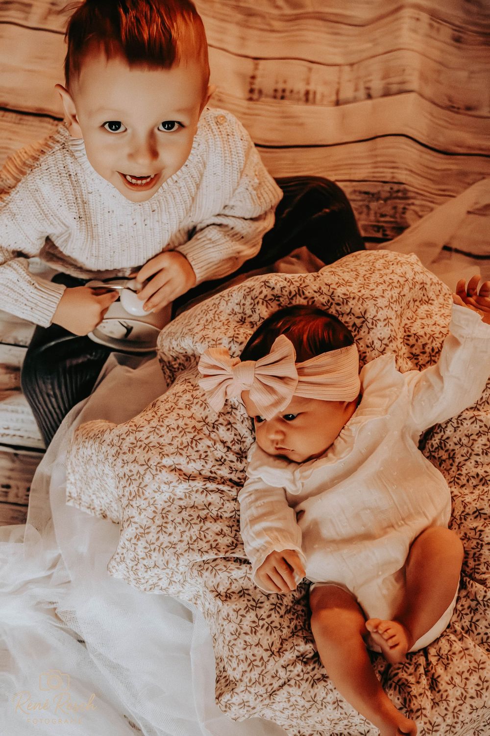 Adorable red-haired toddler boy smiling beside newborn baby girl in white bow headband on floral blanket.