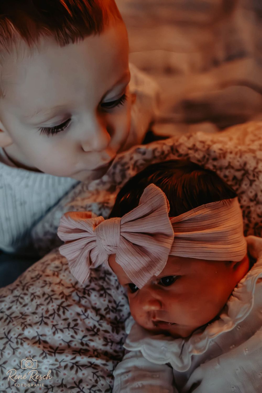 Toddler gazing lovingly at newborn baby sister wearing a pink bow headband, warm golden light photography.