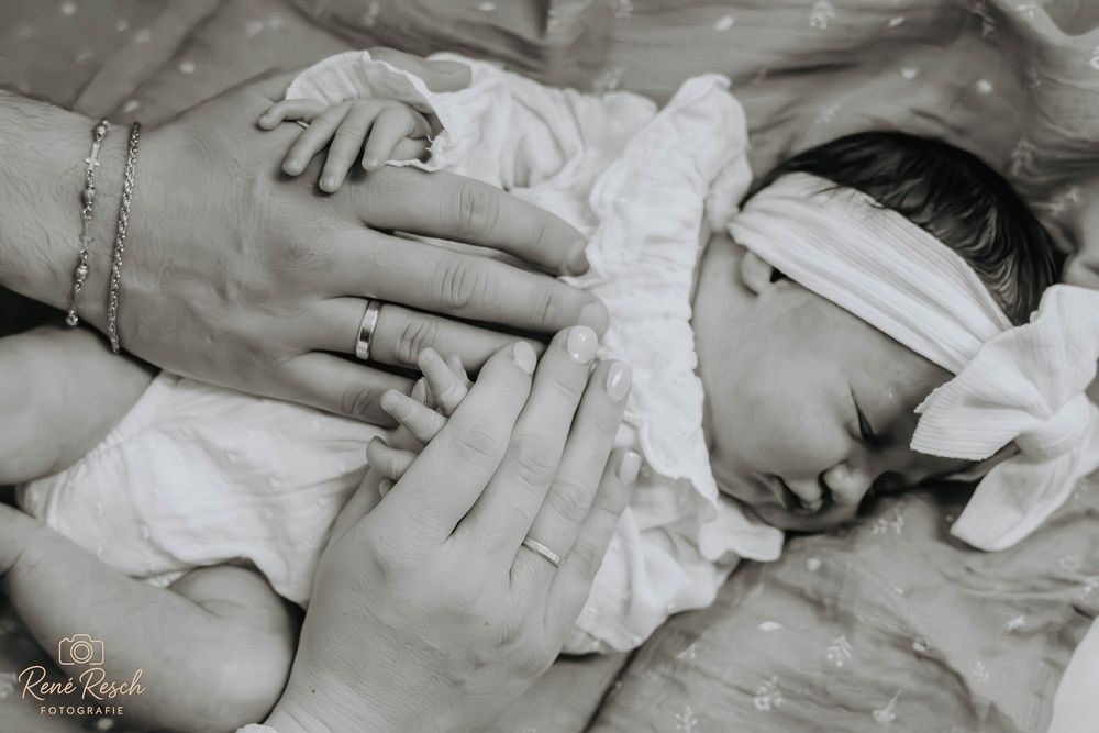 Black and white newborn photo of sleeping baby with parents hands gently cradling their face, wearing white headband.