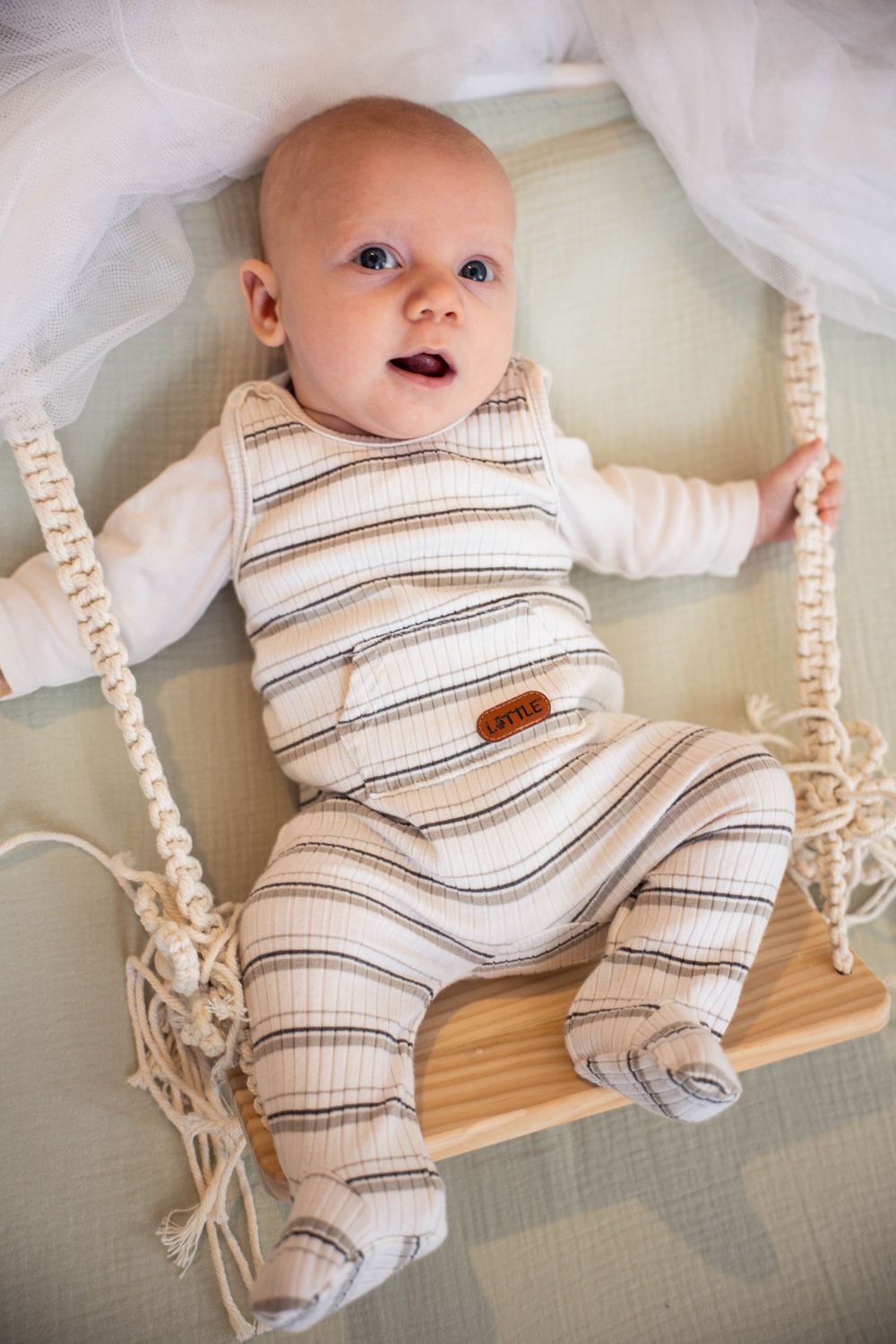 Adorable baby in striped romper sitting on a wooden macrame swing, looking up with bright blue eyes.