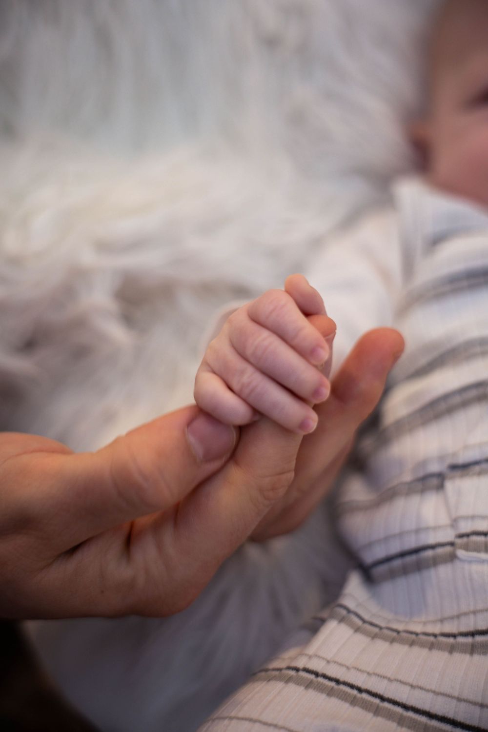 A newborn baby grasping an adult finger while lying on a soft white blanket in a striped onesie.