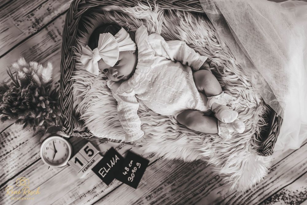 Black and white newborn photo of baby Ellie in a basket with fur, bow headband, clock, and name sign.