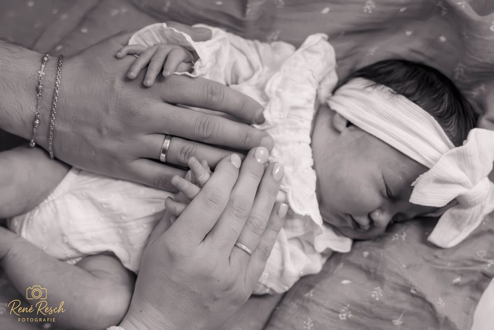 Black and white newborn photo with parents tenderly holding their sleeping baby wrapped in white cloth with a bow headband.