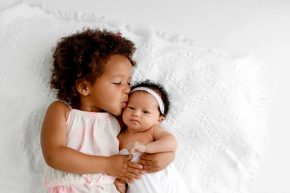 Toddler girl in pink dress kissing her newborn baby sister wrapped in white on a soft white blanket.