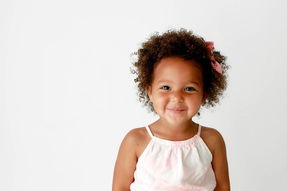 Smiling young girl with curly hair and pink bow wearing a white dress against a light background.
