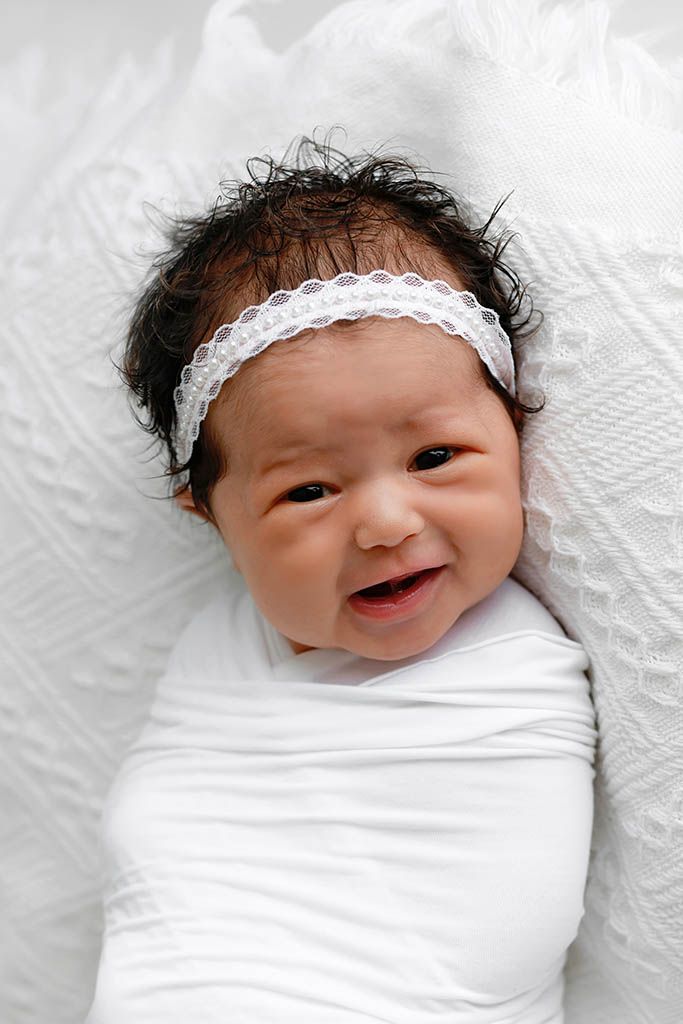 Newborn baby girl swaddled in white blanket wearing a delicate white lace headband, smiling sweetly.