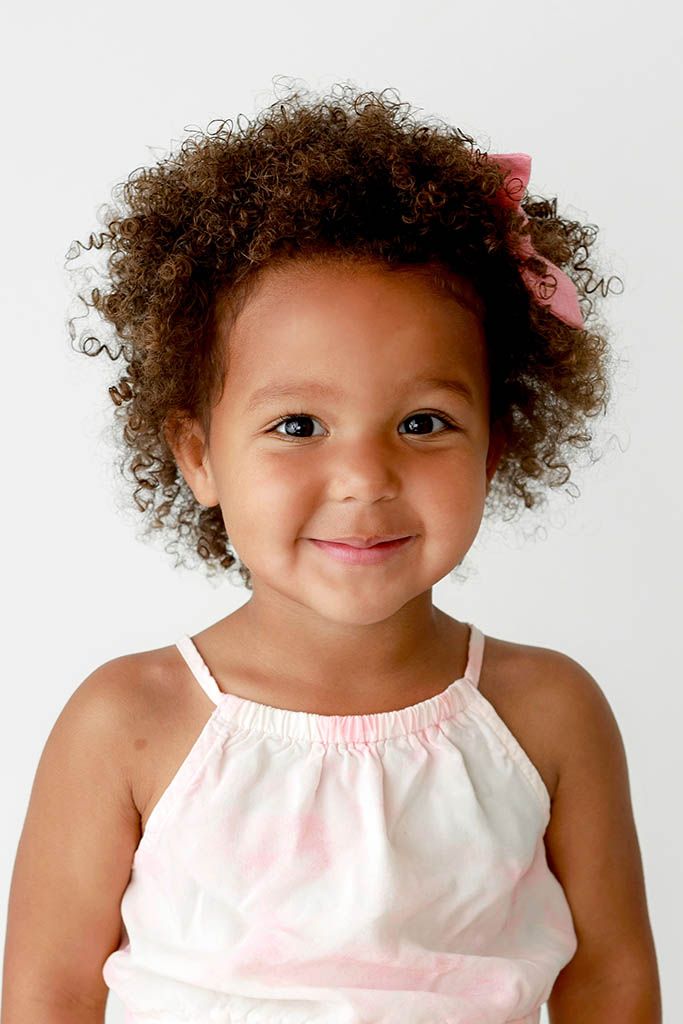 Smiling toddler girl with curly hair and pink bow wearing a white sundress.