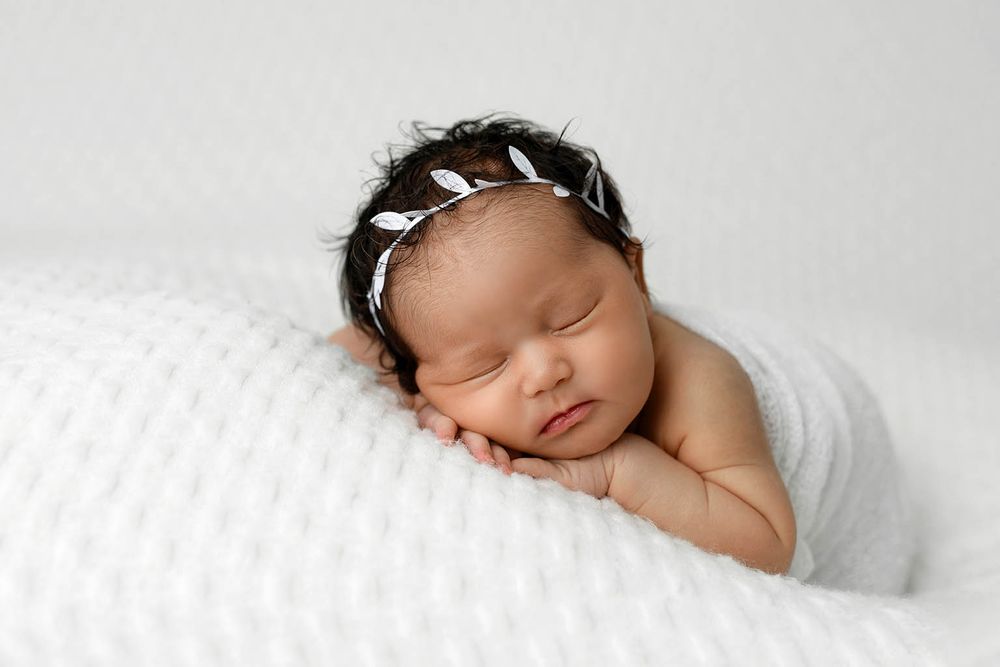 Peaceful newborn baby sleeping on white blanket wearing a delicate floral headband.