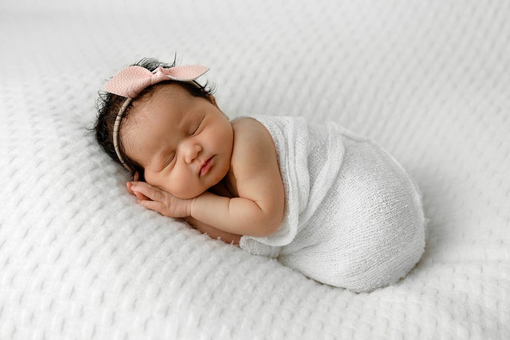 Sleeping newborn baby girl swaddled in white wrap with a pink bow headband on a white textured blanket.