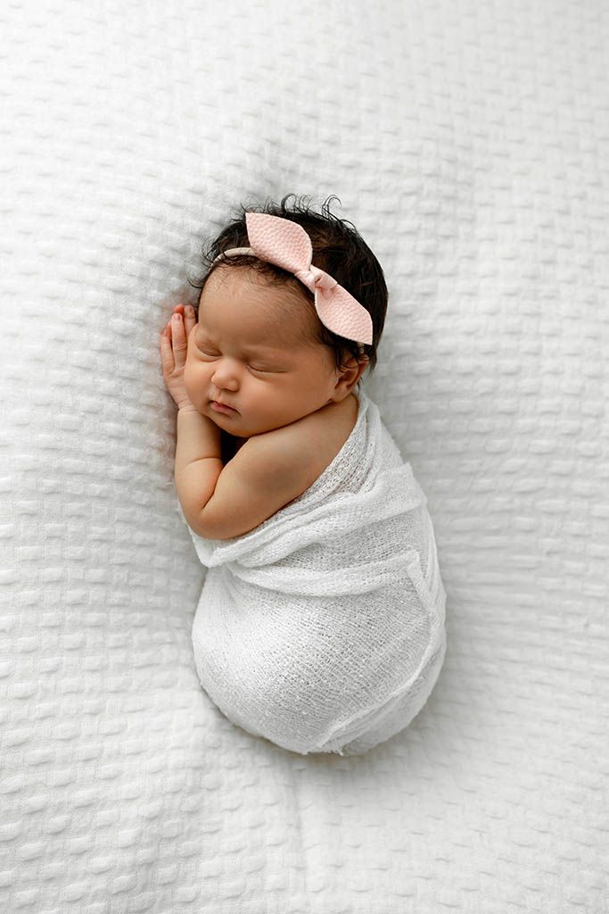 Sleeping newborn baby girl swaddled in white wrap with pink bow headband on white blanket.