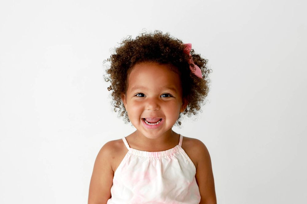 Smiling young girl with curly hair and pink bow wearing a white dress against a light background.