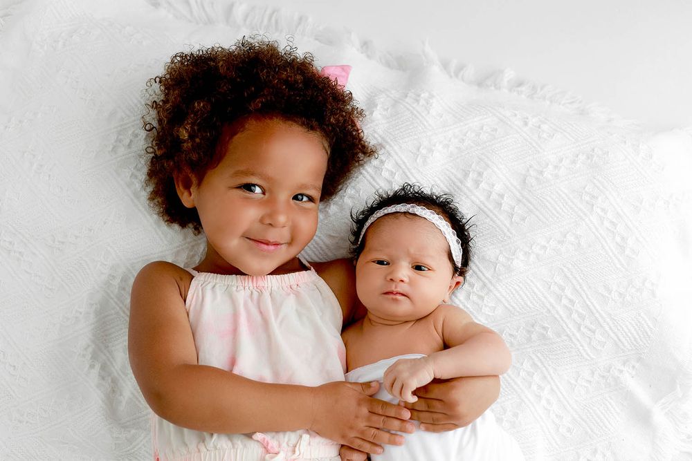A smiling toddler girl in pink dress cuddles a newborn baby with headband on a white blanket.