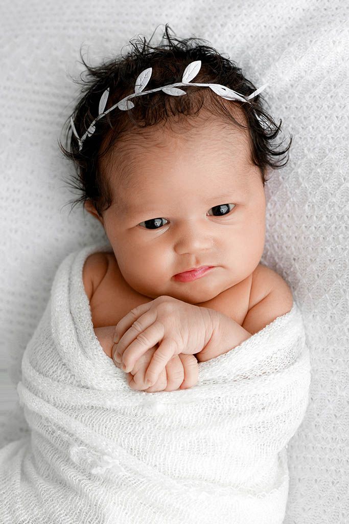 Newborn baby swaddled in white wrap with silver leaf headband, looking up with bright eyes.