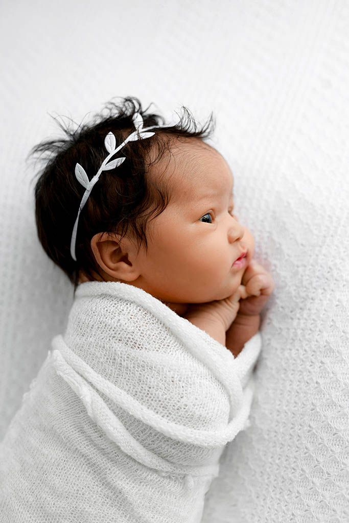 Newborn baby girl swaddled in white wrap wearing a delicate silver leaf headband, lying on a white blanket.