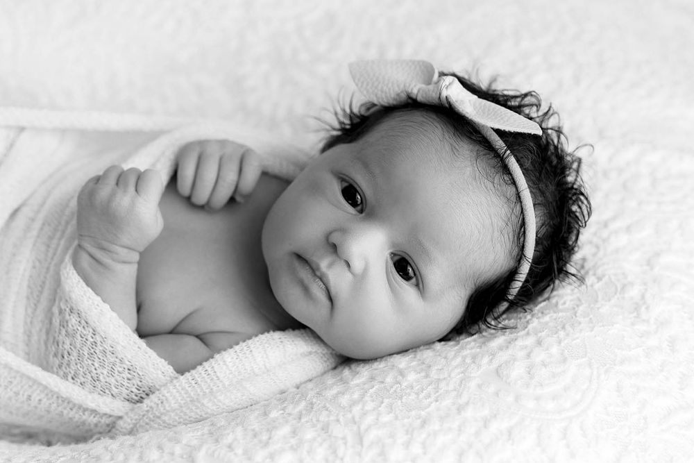 Black and white portrait of an alert newborn baby girl with curly hair wearing a bow headband.