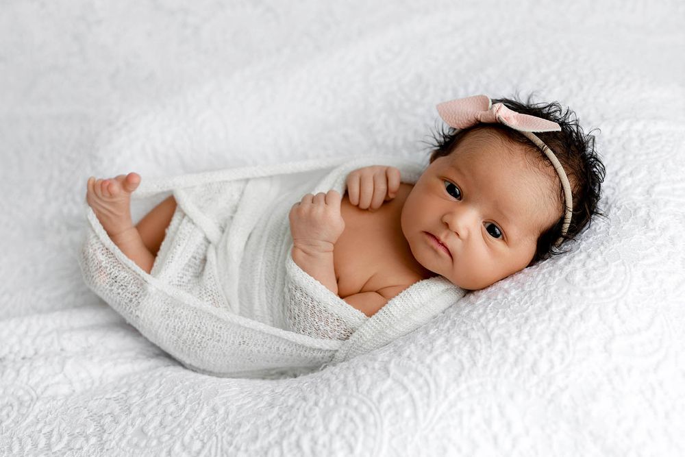 Newborn baby swaddled in white wrap with pink bow headband lying on white textured blanket.