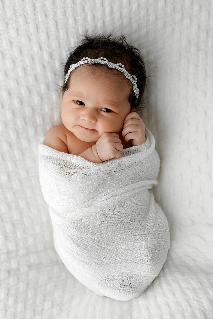 Newborn baby girl swaddled in white wrap with delicate headband, smiling softly while lying on white textured blanket.